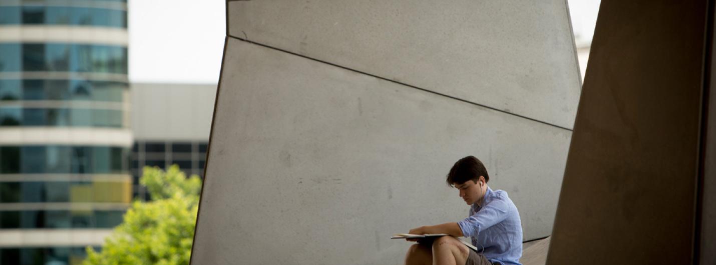 a male student sits outside on the concrete ledge outside Gates Hall in summer