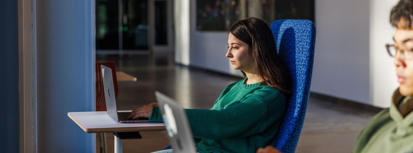 a female student with brown hair sits and works on her laptop in the hallway