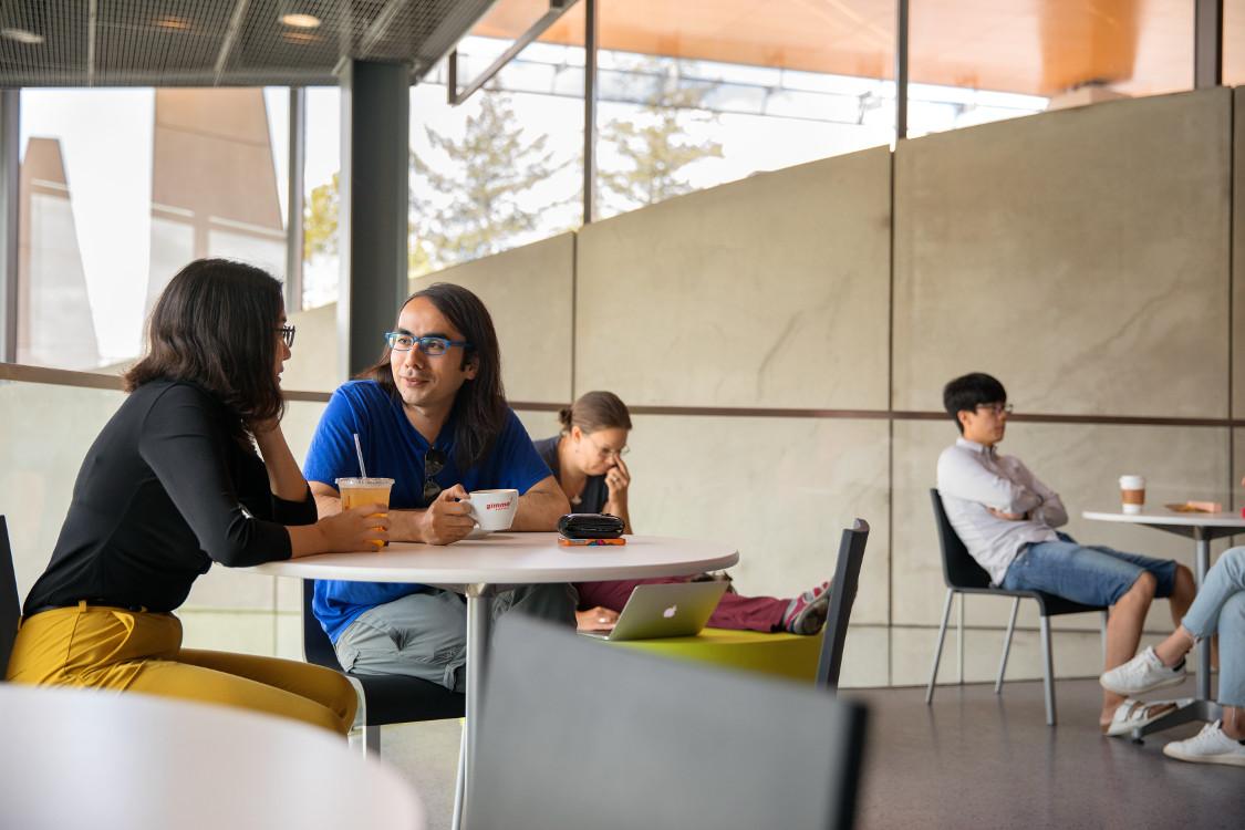 a male and female sit at a table with coffee outside with other people sitting at tables in the background