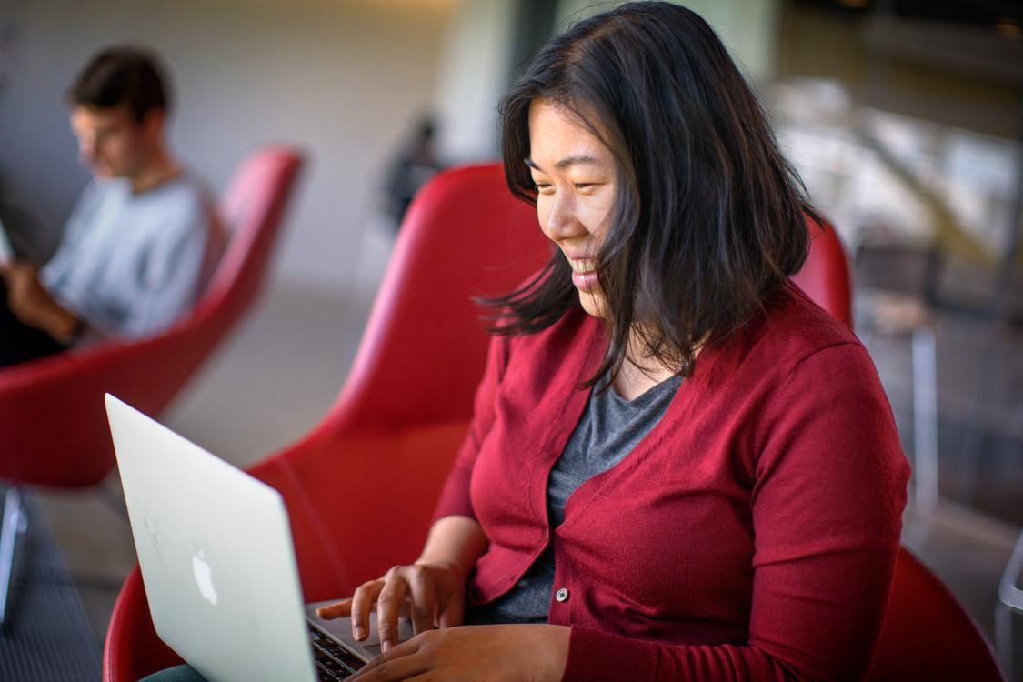 A woman sits in a red chair. She is smiling while working on a laptop and wearing a red cardigan.